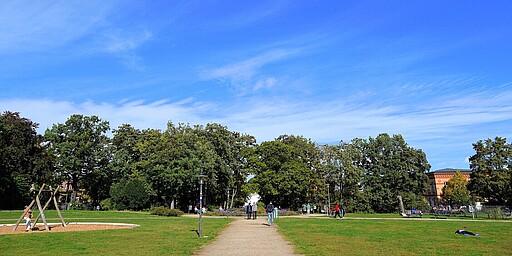 Spazierweg im Forckenbeckplatz bei strahlendem Himmel Ein breiter Kiesweg führt durch den Forckenbeckplatz. Links sieht man eine Holzschaukel auf einem Kinderspielplatz, und rechts erstrecken sich grüne Wiesen. Mehrere Menschen sind im Park zu sehen, einige stehen und unterhalten sich, andere spazieren. Am Horizont erstreckt sich ein blauer Himmel mit leichten, weißen Wolkenstreifen.