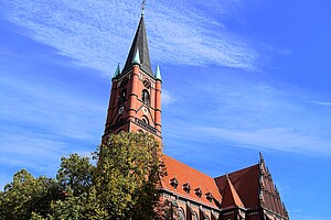 Evangelische Samariterkirche im Samariterviertel Die beeindruckende rote Backsteinkirche mit einem hohen Kirchturm, der in den blauen Himmel ragt. Der Turm ist mit grünen Spitzen und Ornamenten geschmückt. Im Vordergrund befindet sich eine Baumreihe, deren Blätter teilweise das Gebäude verdecken.