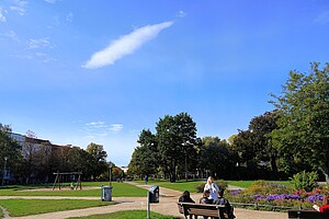 Ruhiger Parktag auf dem Forckenbeckplatz Eine Bank steht auf einem Kiesweg im Park, auf der zwei Menschen sitzen und plaudern. Im Hintergrund sieht man grüne Wiesen und blühende Blumenbeete. Einige Bäume umrahmen den Park, und der Himmel ist klar und blau, bis auf eine große, längliche Wolke.
