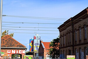 Graffiti und Straßenbahn in Friedrichshain Ein urbaner Berliner Ausblick mit Graffiti-bedeckten Backsteinmauern im Vordergrund. Eine Straßenbahnhaltestelle ist zu erkennen, und im Hintergrund erhebt sich ein modernes Hochhaus mit bunter Fassadenkunst. Oberleitungen der Straßenbahn durchkreuzen den Himmel.