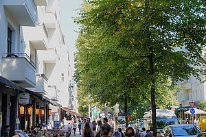 Simon-Dach-Straße bei Tageslicht Eine belebte Berliner Straße, gesäumt von Altbauhäusern mit weißen Fassaden. Links im Bild befinden sich Restaurants und Cafés mit Außenbereichen, in denen viele Menschen sitzen. Rechts steht eine dichte Baumreihe, die Schatten auf die Straße wirft. Fußgänger und Radfahrer beleben die Szene.